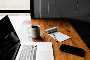 A mobile phone, notepad, cup of coffee and laptop sprawled across a wooden table.