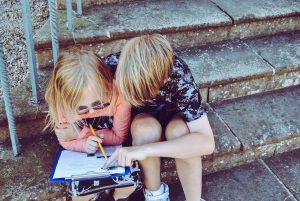 Two children sitting on cement steps examine a paper on a clipboard.
