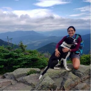 Brown haired young woman with dog smiling on top of mountain