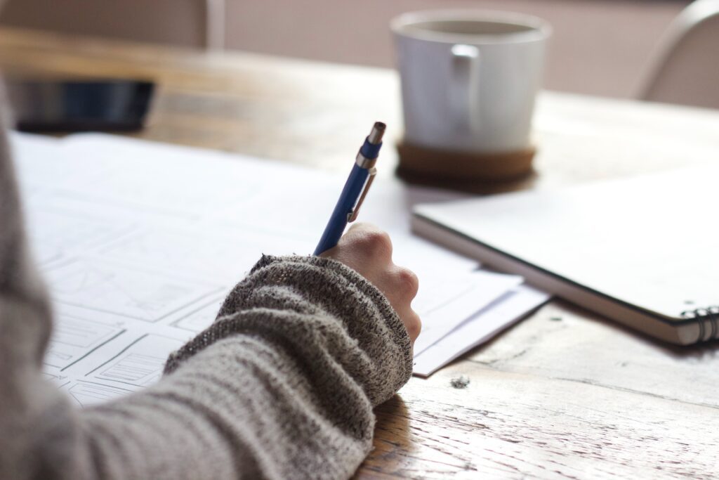 person working at a table, writing on a notebook, with a cup of coffee in the background