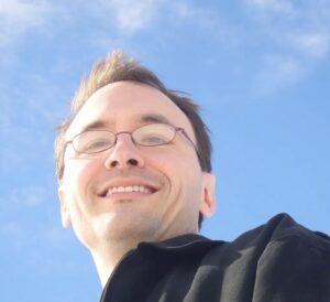 white male-presenting person with short hair and glasses, smiling