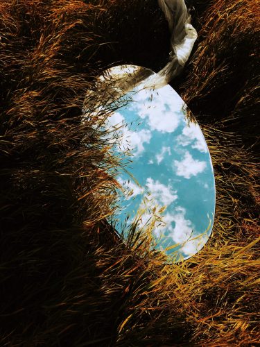 a mirror laying in the grass with a cloudy blue sky reflected in the glass